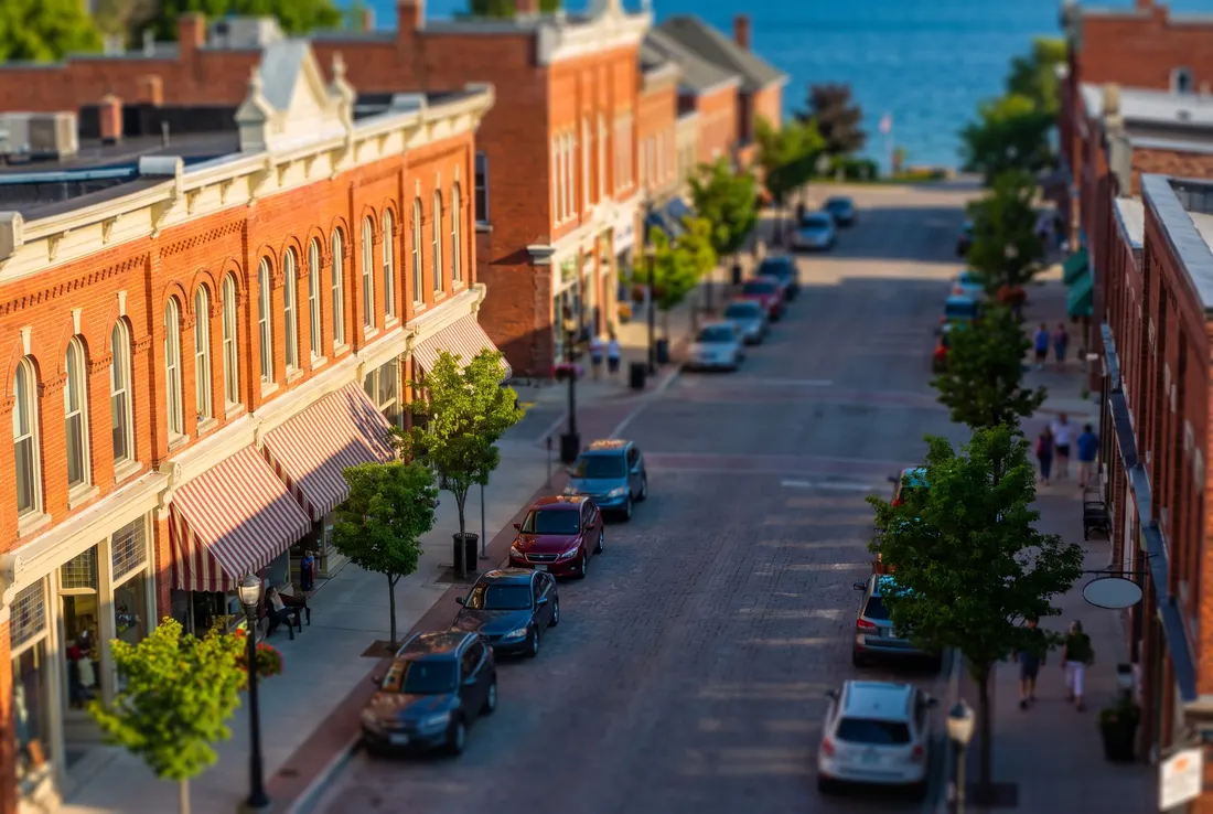 Tilt-shift miniature of a heritage main street with red-brick shopfronts, striped awnings and a glimpse of a blue lake